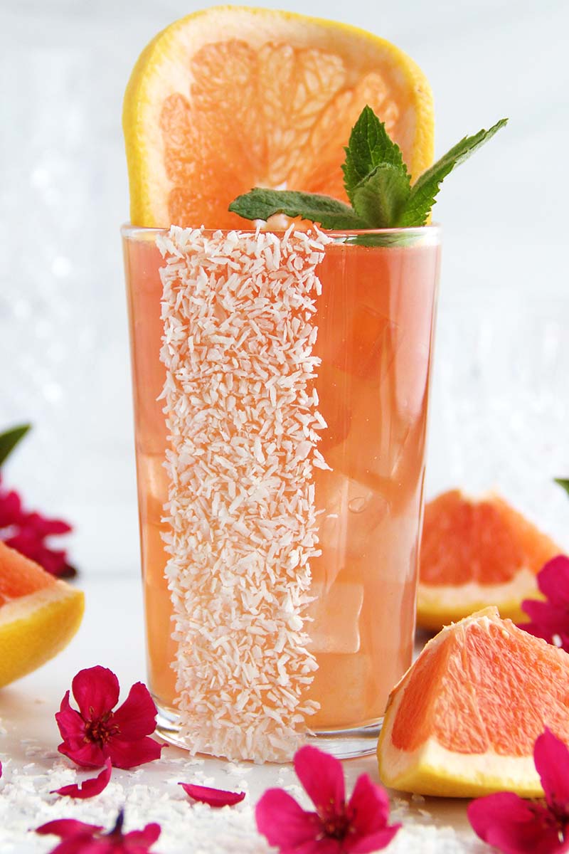 A tropical Coconut Paloma cocktail, shown in a tall serving glass garnished with a grapefruit slice, fresh mint, and shredded coconut.