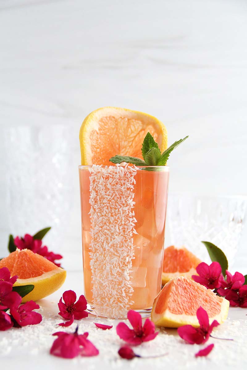 Pink-colored Coconut Paloma, shown against a white background with pink flowers and grapefruit wedges.