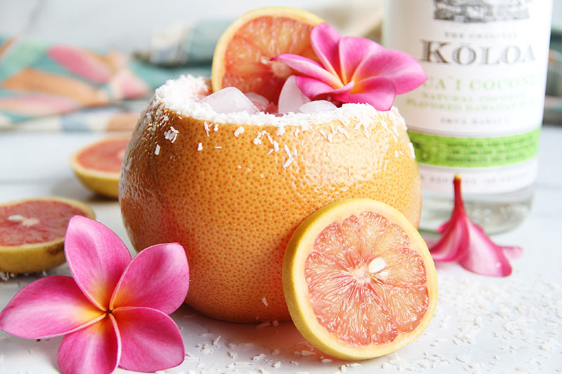 Coconut Paloma cocktail, served in a hollowed-out grapefruit, shown with pink tropical flowers and a bottle of coconut rum.