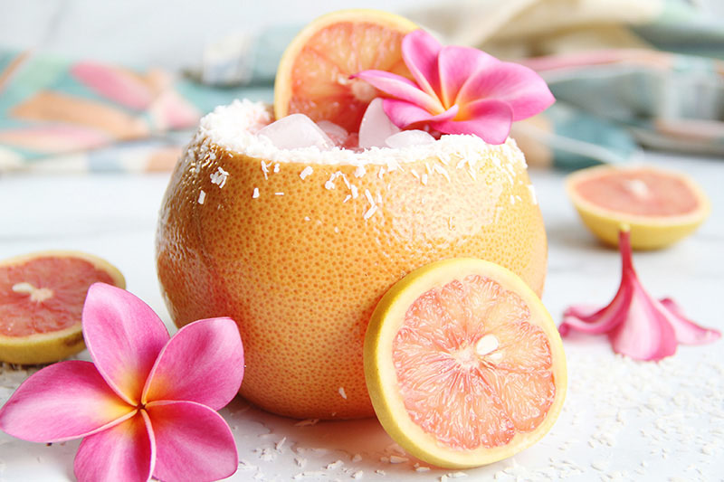 Coconut Paloma cocktail, shown in a hollowed-out grapefruit with pink flowers and grapefruit wedges.