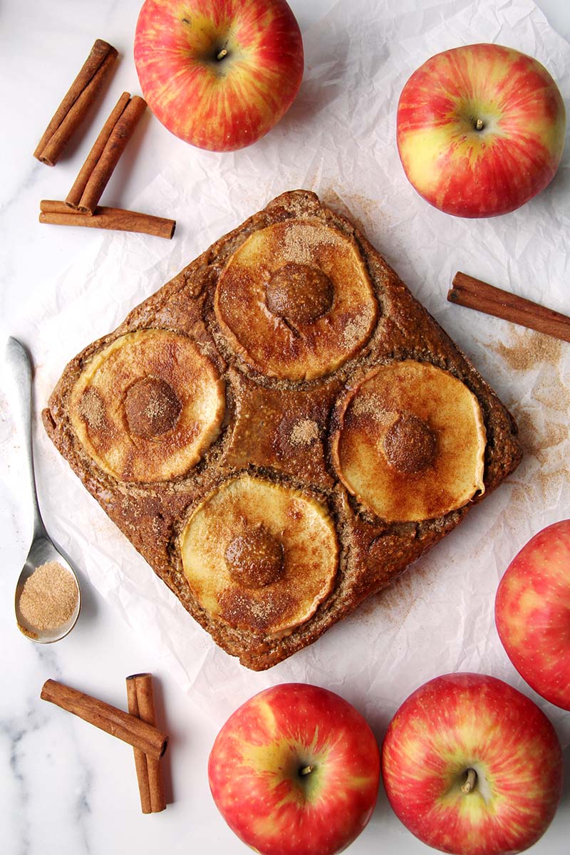 Gluten-free apple cider donut cake, shown on a white surface with apples and cinnamon sticks.