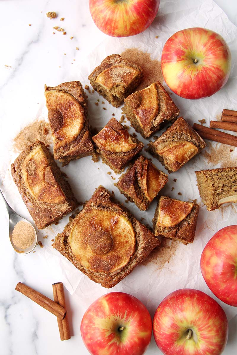 Apple cider donut cake slices, shown from above on a white surface with honeycrip apples, cinnamon sticks, and cinnamon sugar.