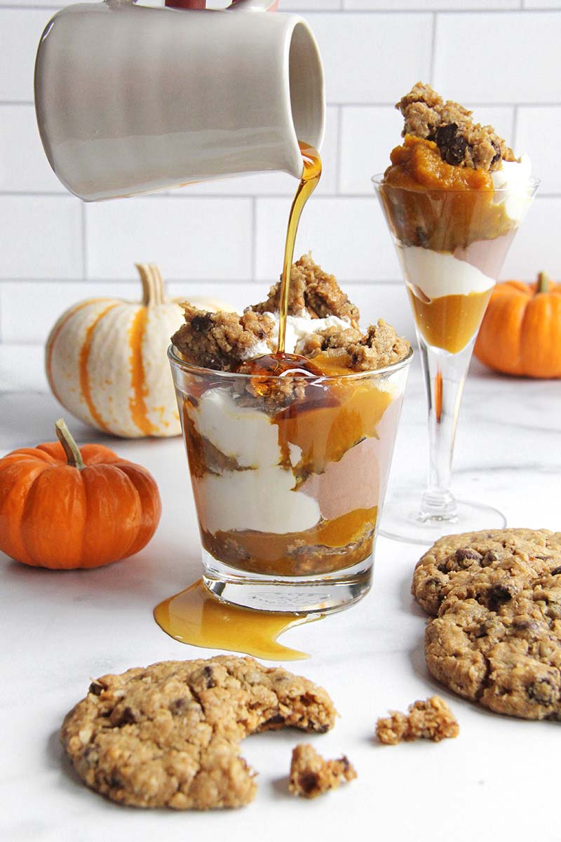 Woman's hand drizzling maple syrup from a pitcher on top of a pumpkin cookie trifle in a serving glass.