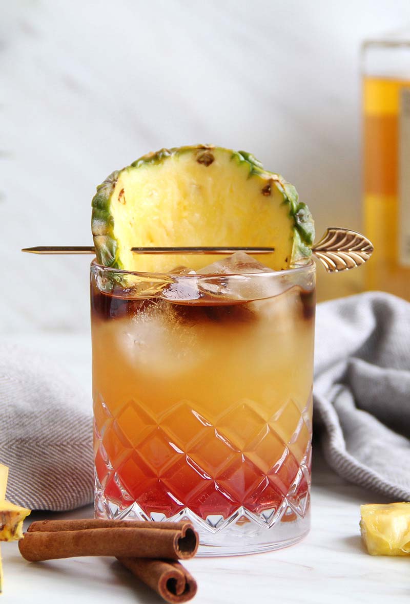 Chai Tai cocktail with fall colors (red, yellow, brown), shown with cinnamon sticks and a bottle of rum.