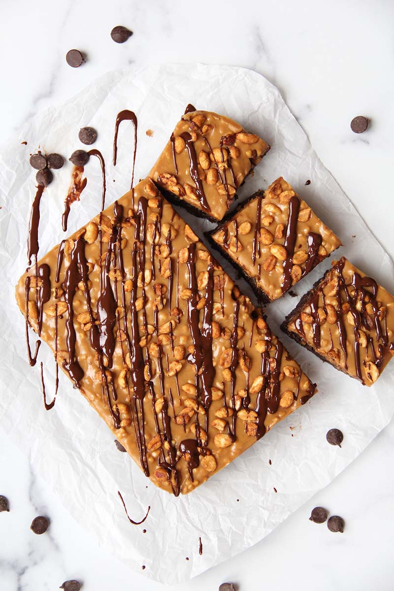 Healthy vegan Snickers brownies on a white kitchen counter, shown with drizzled chocolate and dark chocolate chips.
