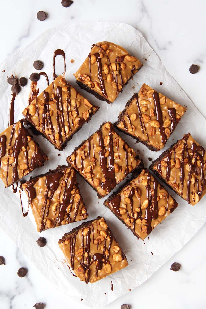 Vegan Snickers brownies on a white kitchen counter, shown with freshly-drizzled chocolate and chocolate chips.