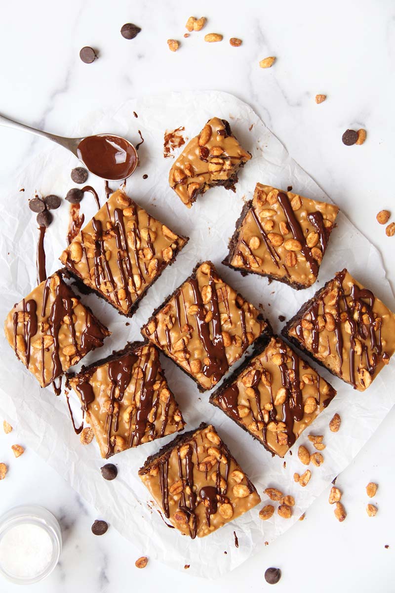 Healthy Snickers brownies shown with a messy chocolate-covered spoon, extra chocolate chips, and extra peanuts.