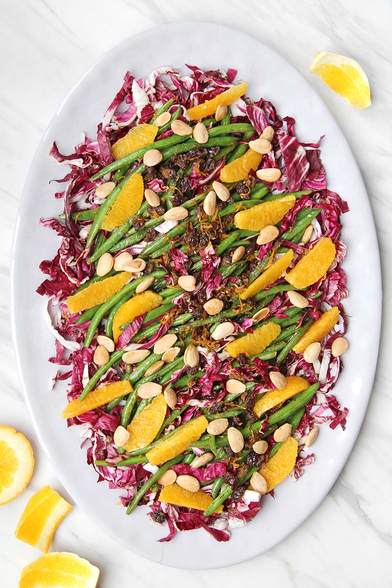 Large serving platter of colorful Spanish green beans on a white surface, shown next to orange peels.