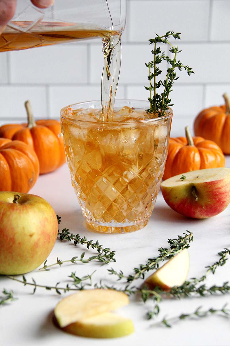 Hand pouring a fall Old Fashioned variation from a cocktail stirring glass into a serving glass filled with ice and a thyme sprig.