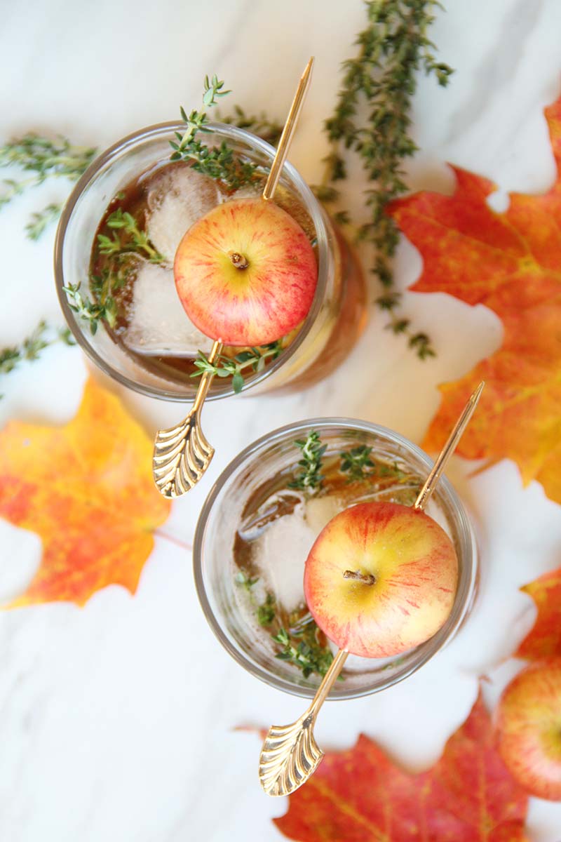 Overhead view of two Apple Thyme Old Fashioned cocktails, shown on a white surface with thyme sprigs and orange maple leaves.