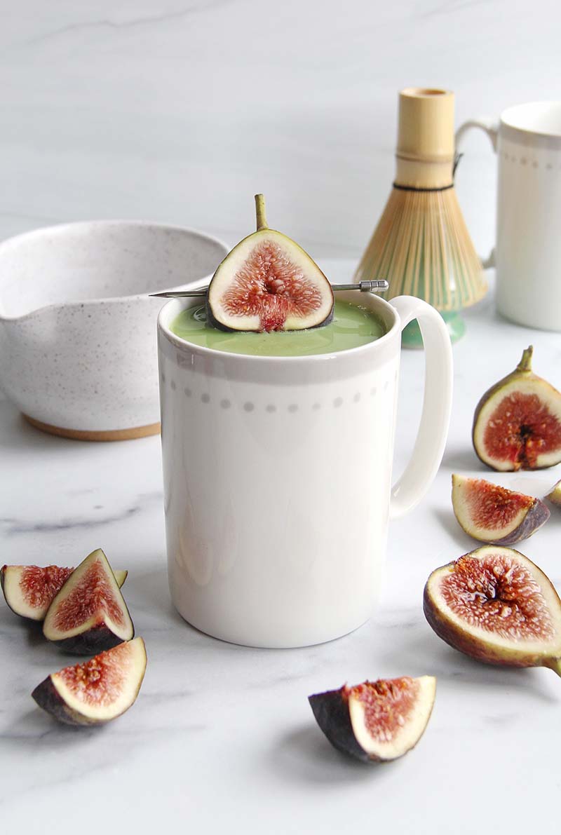 Honey fig matcha latte in a white kitchen, showing the green matcha and red fig garnish, placed next to white mugs and a matcha whisk.