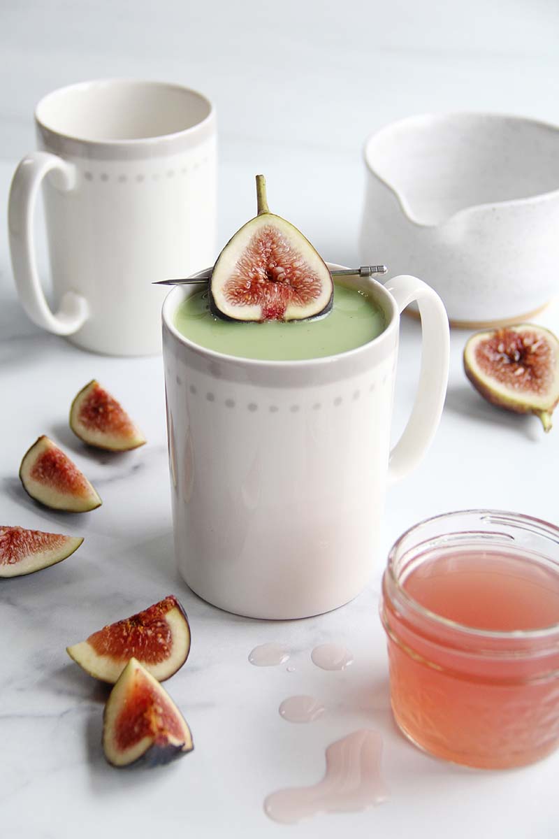 White mug of honey fig matcha latte on a clean white background, topped with a fresh fig half and surrounded by scattered fig slices, as well as a jar of honey fig syrup in the foreground.