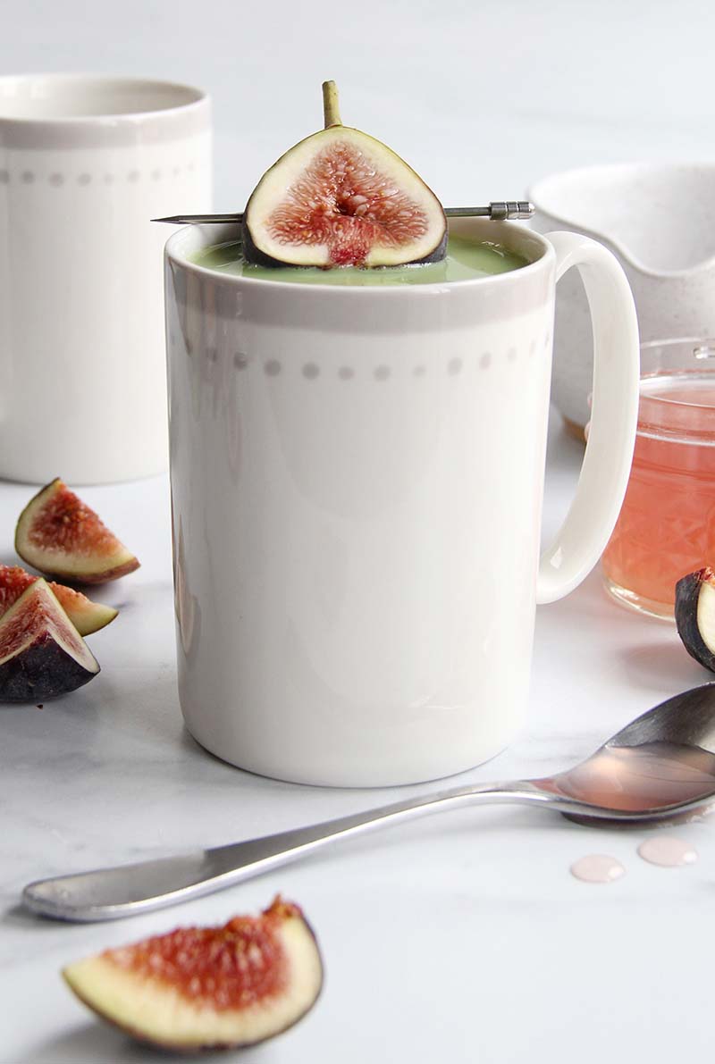 White porcelain mug filled with honey fig matcha latte, styled with a fresh fig half on top and assorted fig pieces arranged on a white kitchen surface.