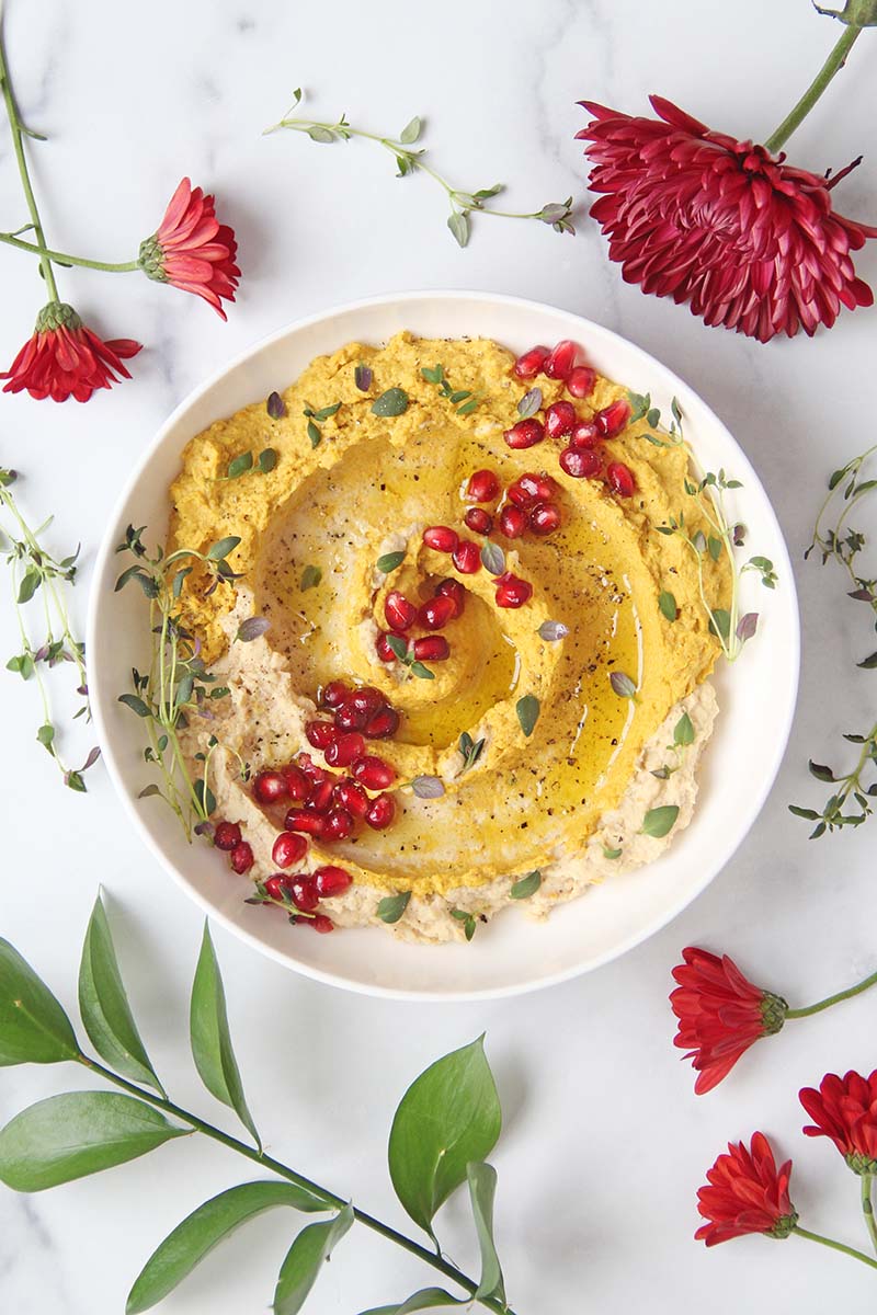 Sweet potato hummus in a white serving bowl on a white marble surface, shown with red holiday flowers.