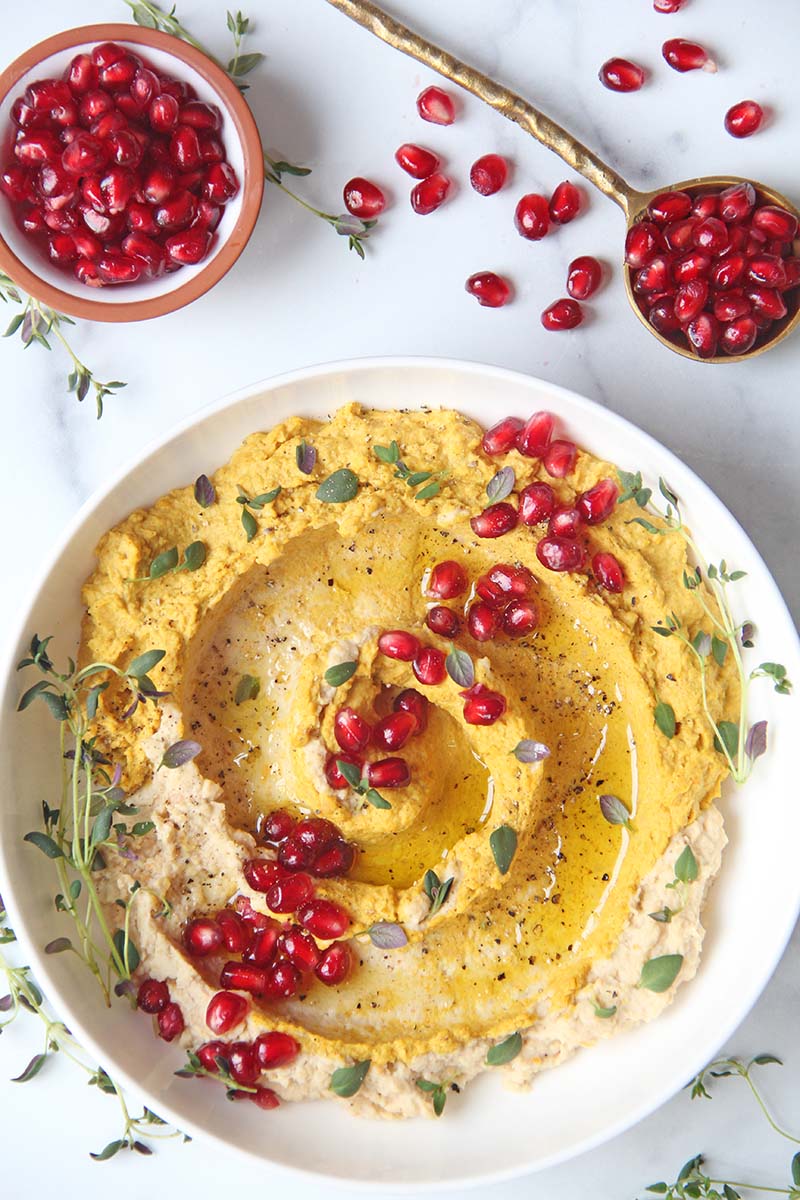 Overhead view of holiday sweet potato hummus, shown with a small bowl and spoonful of pomegranate arils.