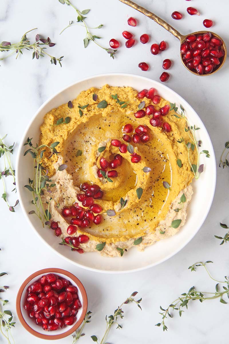 Holiday sweet potato hummus, shown on a white marble surface next to pomegranate arils and fresh thyme sprigs.
