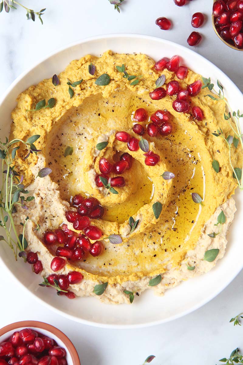 Cose-up of the surface of a bowl of sweet potato hummus garnished with a drizzle of olive oil, pomegranate arils, and thyme leaves.