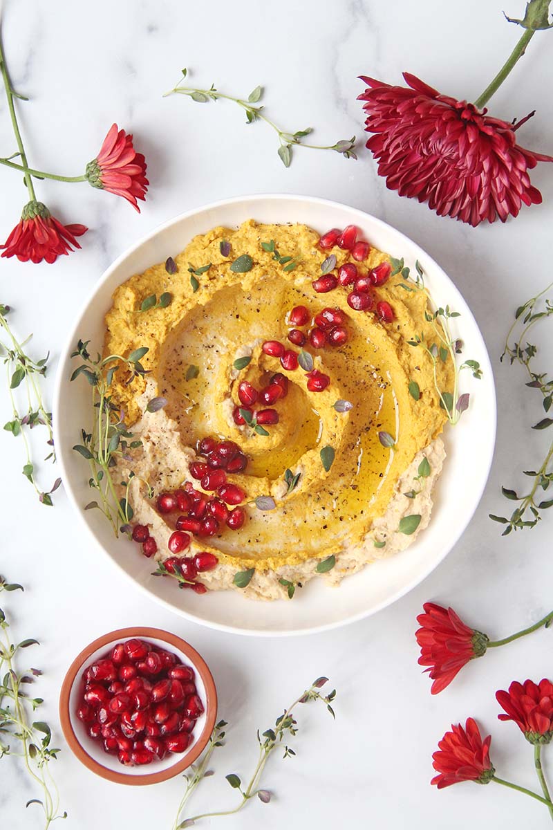 Bowl of orange-colored sweet potato hummus shown next to red holiday flowers and fresh thyme.