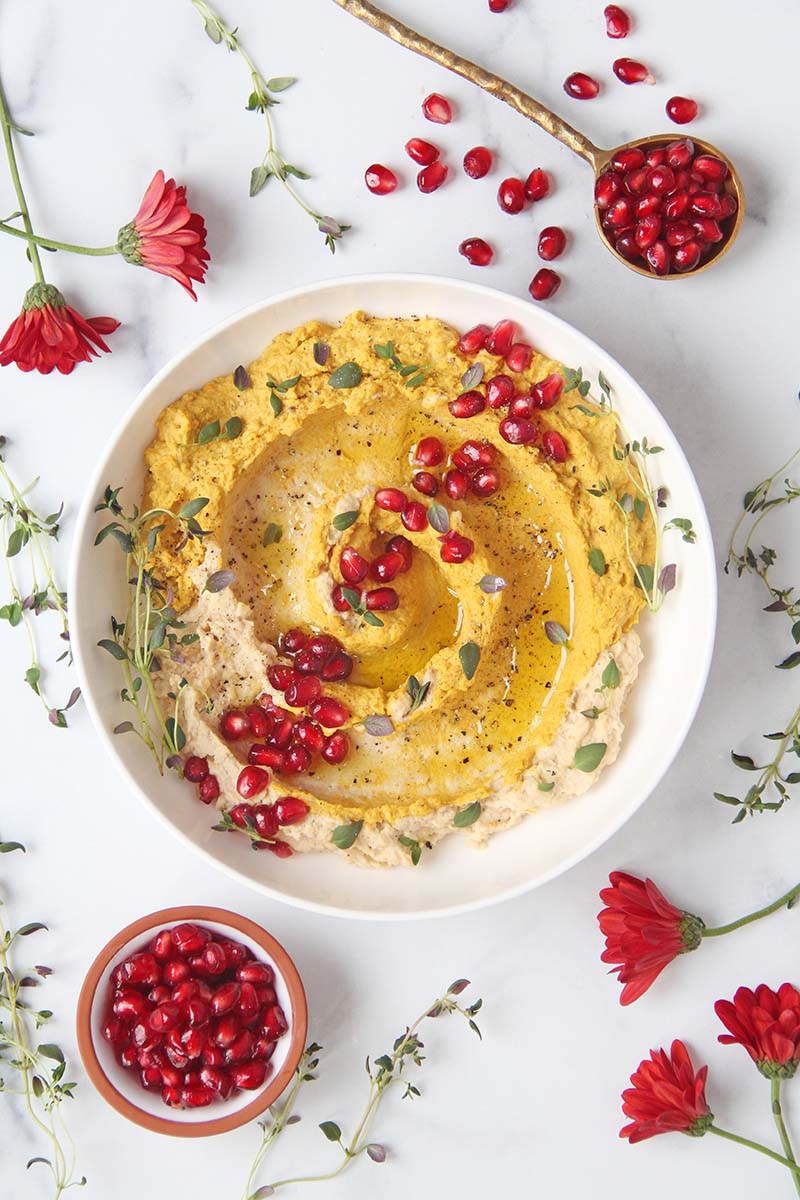 Overhead view of a bowl of sweet potato hummus, shown next to a small bowl and a spoon full of pomegranate arils.