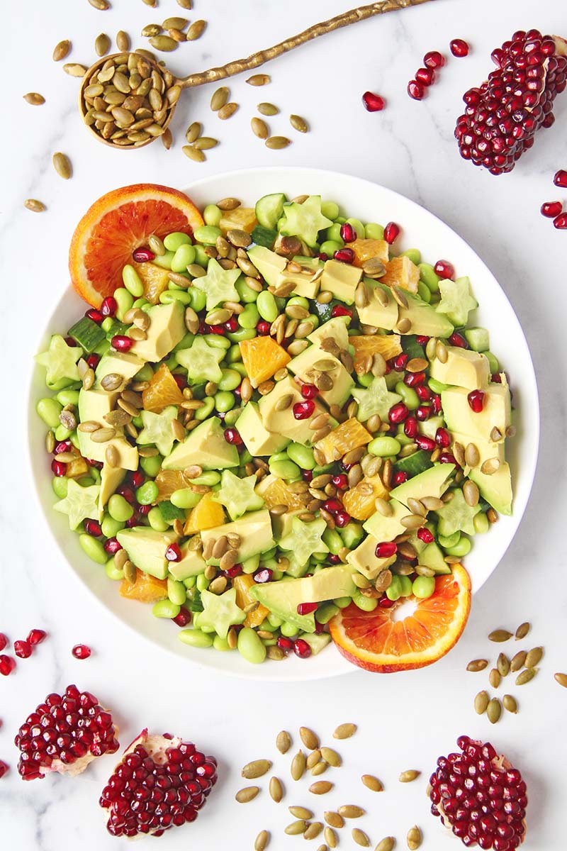 Vibrant, healthy holiday edamame salad with cucumbers, avocado, orange, and pomegranate, shown in a white serving bowl.