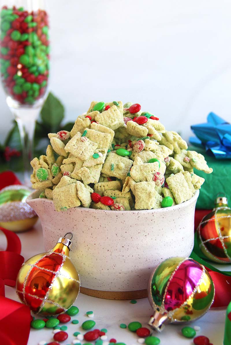 Christmas matcha puppy chow in a white bowl, surrounded by festive holiday ornaments and topped with red and green sprinkles and chocolate candies.