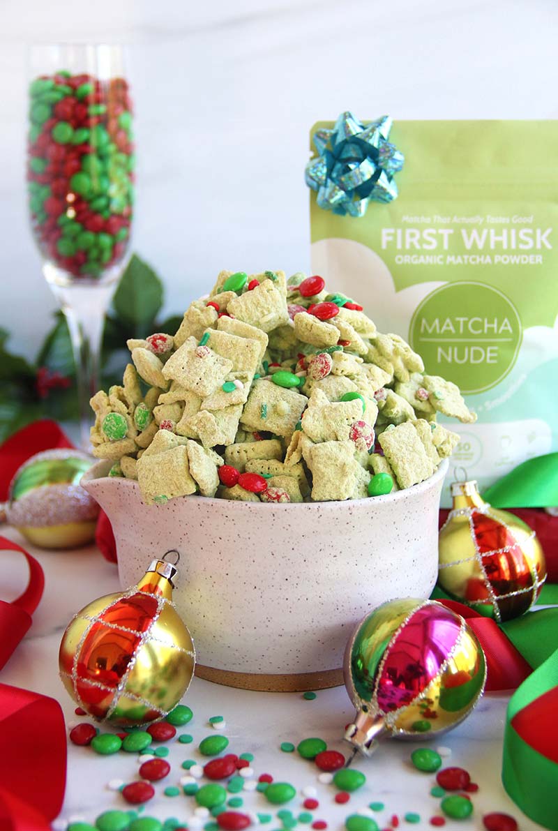 Vibrant green Christmas matcha puppy chow in a white bowl with red and green sprinkles and chocolate candies, styled with holiday decorations and a bag of matcha powder in the background.