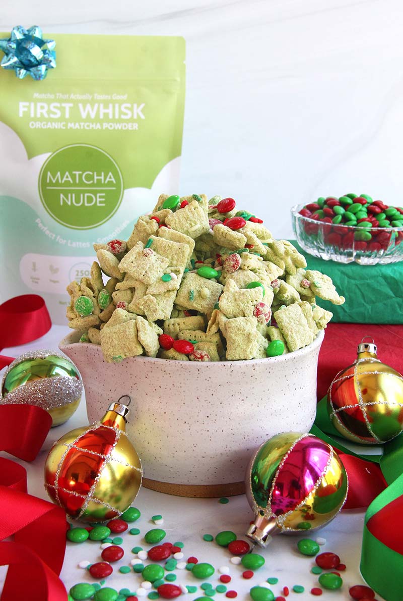 Festive matcha puppy chow made with white chocolate and matcha, served in a white bowl with Christmas ornaments and a matcha bag in the background.