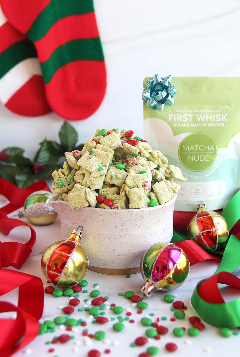 Easy Christmas matcha puppy chow in a white bowl, styled with holiday ornaments, Christmas stockings, and a bag of matcha behind the bowl.