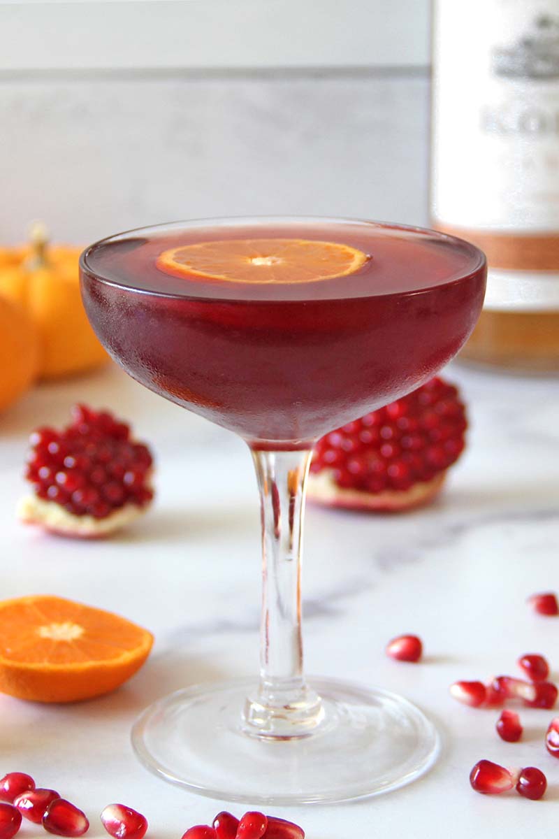 Spiced pomegranate daiquiri in a coupe glass garnished with a clementine slice, shown with fresh pomegranate arils and a bottle of rum in the background.