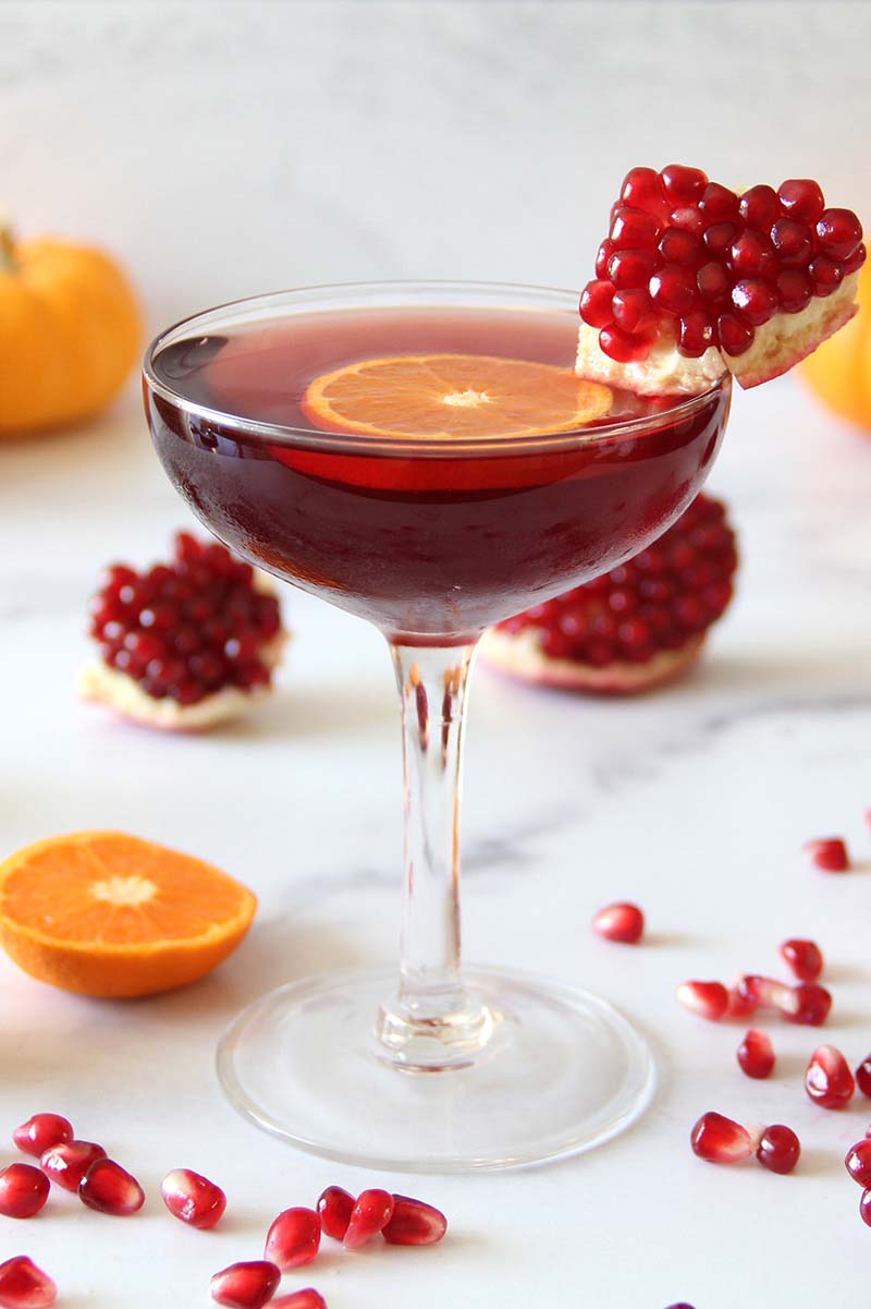Festive spiced pomegranate daiquiri in a coupe glass, garnished with a piece of pomegranate and a clementine slice, surrounded by more fresh pomegranate and citrus.