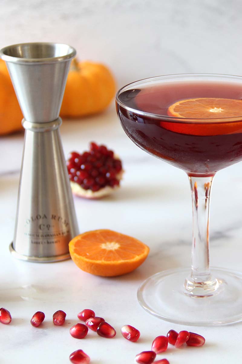 Festive holiday spiced pomegranate daiquiri with garnished with a clementine slice, shown with a measuring jigger and seasonal ingredients.