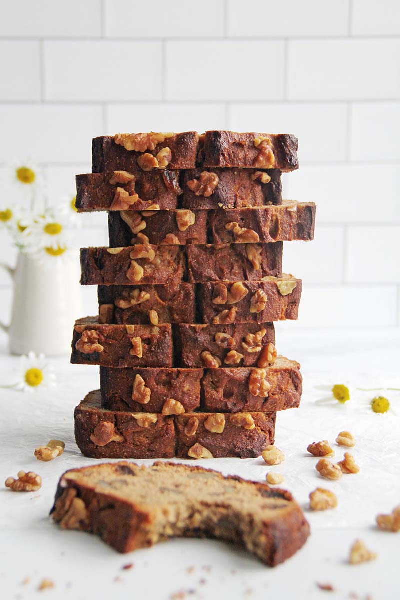 Vertical stack of walnut-topped gluten-free chickpea flour banana bread slices, with one slice in the foreground that a bite has been taken from.