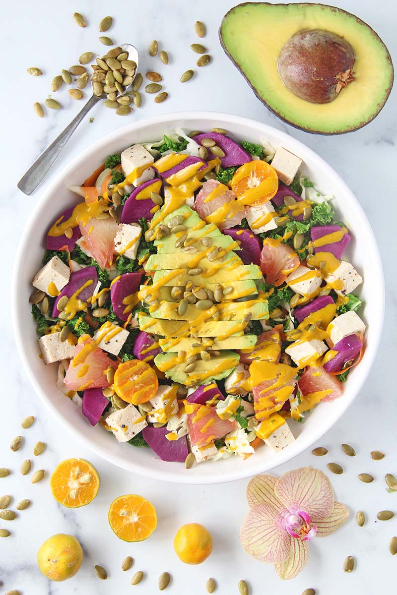 Creamy turmeric tahini dressing drizzled over a colorful rainbow salad with kale, purple sweet potato, tofu, and avocado.