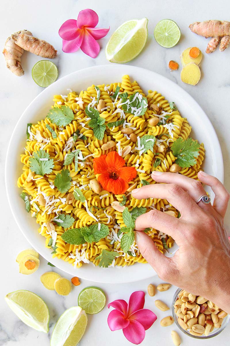 Serving dish of vegan turmeric pasta garnished with peanuts, fresh cilantro, mint, and shredded coconut shown alongside lime wedges and fresh ginger.