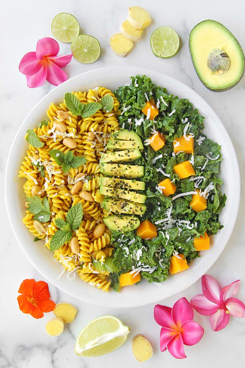 Healthy vegan turmeric pasta portioned for dinner, served with a kale salad and avocado, shown next to lime and ginger root slices.