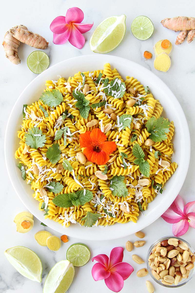 Creamy turmeric coconut milk pasta in a white serving bowl, topped with peanuts, fresh herbs, and shredded coconut, shown next to lime edges, turmeric root, and ginger root.