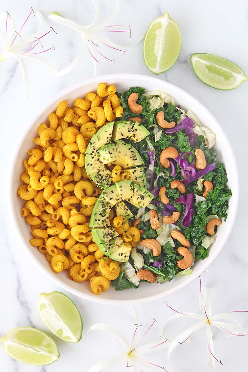 Plate of vegan turmeric pasta with a side salad, photographed on awhite background with lime wedges and tropical flowers.