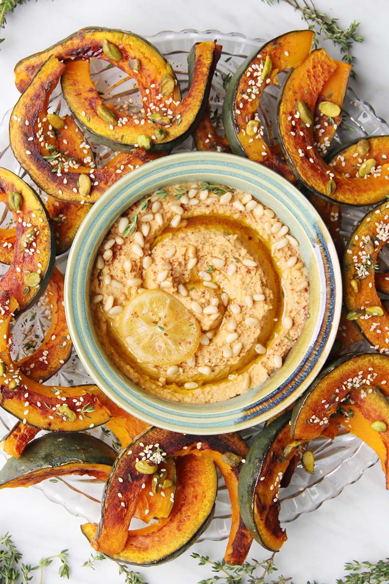 Roasted kabocha squash dippers seasoned with sumac and thyme, topped with sesame seeds and pistachios, served with lemon hummus, all presented on a glass serving platter.