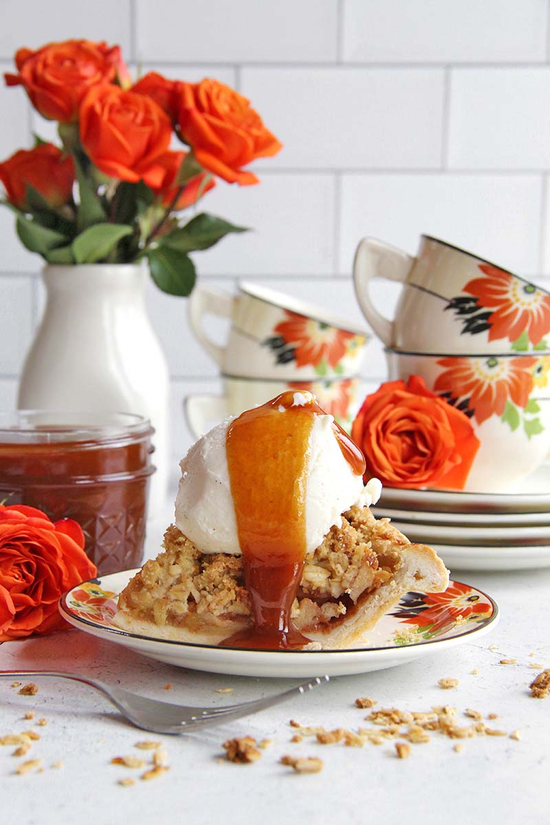 Slice of apple pie topped with vanilla ice cream and rum caramel sauce, with caramel dripping onto the plate, shown in a white kitchen.
