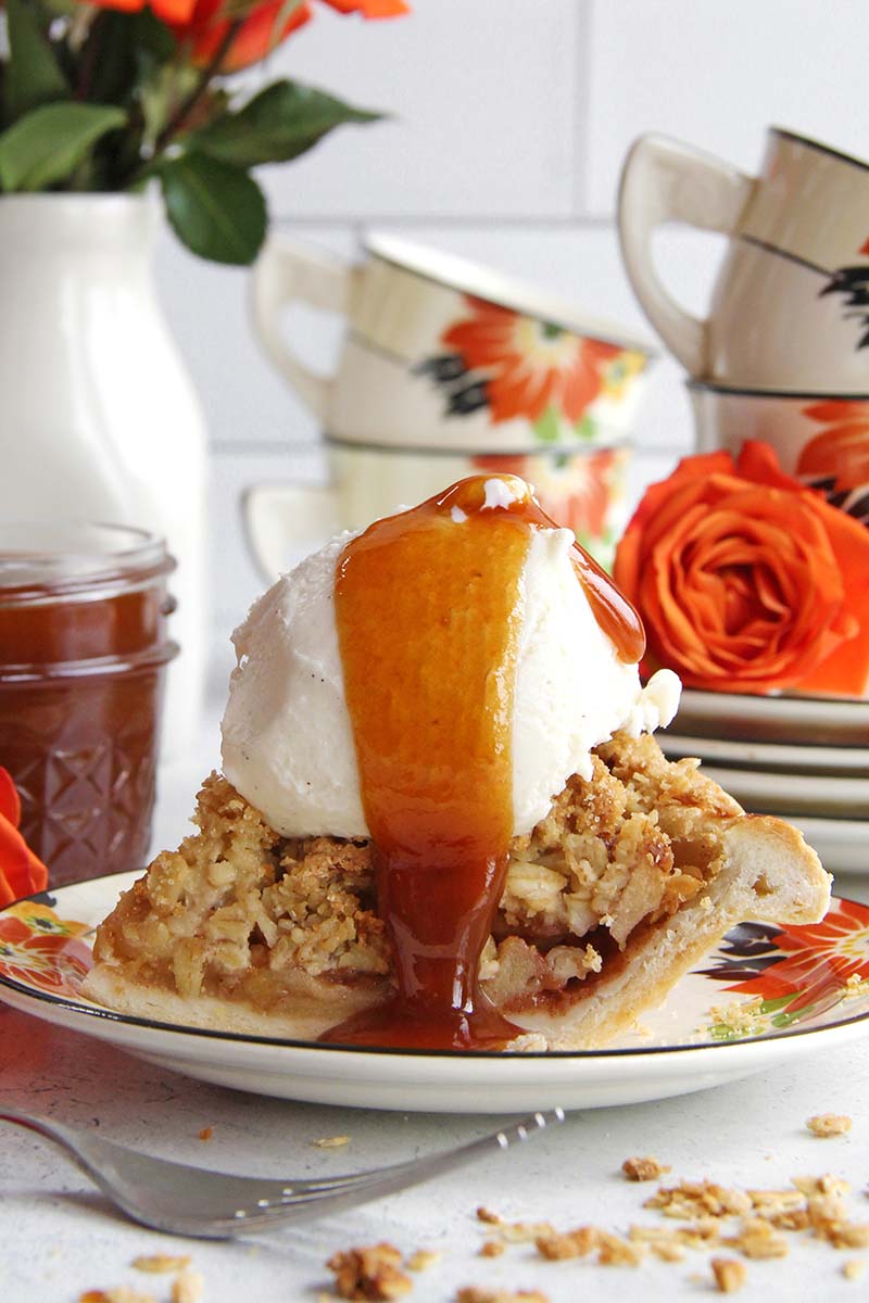 Close-up of a slice of apple pie topped with vanilla ice cream and rum caramel sauce, shown in a white kitchen with fall flowers and a jar of caramel in the background.