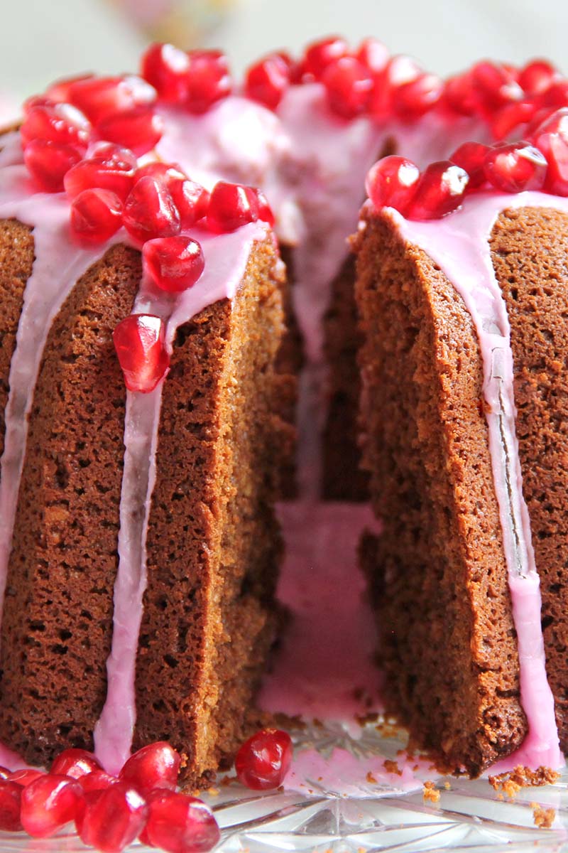 Close-up side view of a gingerbread blender cake with pink icing and pomegranate arils, showing a single slice cut from the cake.