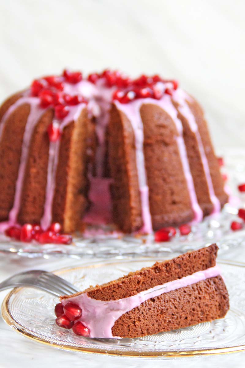 Gingerbread blender bundt cake with pink icing and pomegranate arils, with a sliced piece served on a plate in front of  the cake.