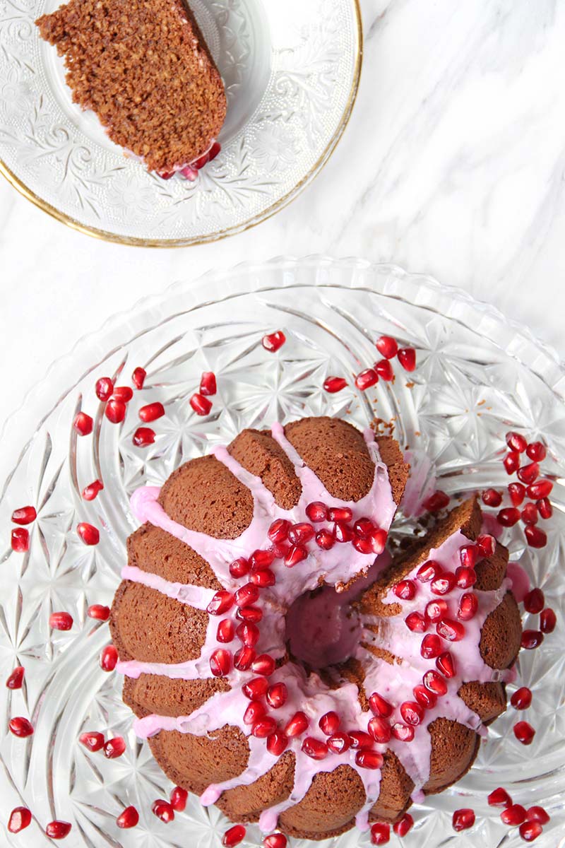 Overhead view of a gingerbread blender bundt cake with pink icing and pomegranate arils, with one slice plated next to the cake.