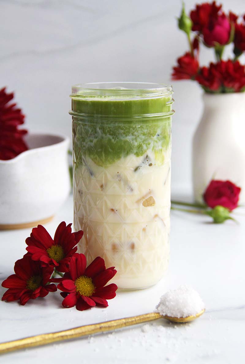 Iced salted maple matcha latte in a glass jar, shown with flaky sea salt in a spoon in the foreground, set in a white kitchen.