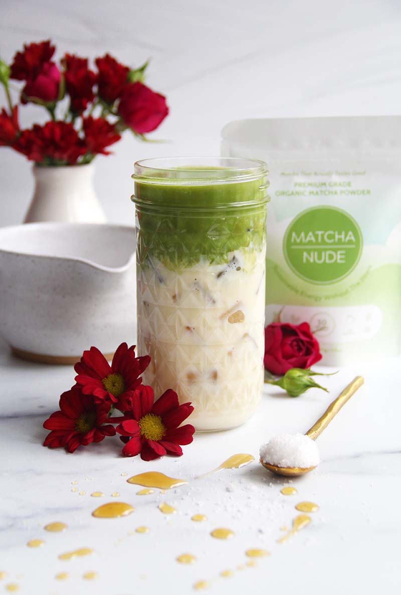 Iced salted maple matcha latte in a glass jar, shown with a matcha powder bag in the background and drops of maple syrup visible on the marble countertop in the foreground.