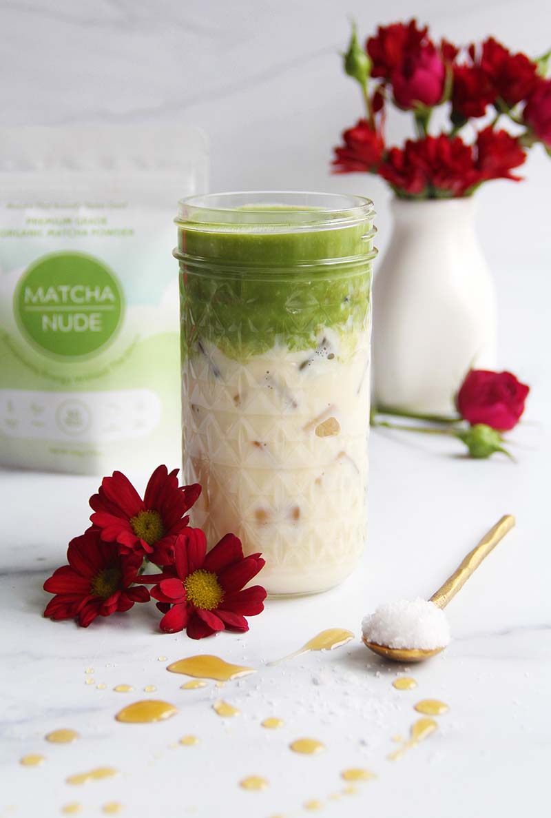 Tall glass jar of iced maple matcha latte with sea salt, shown with maple syrup droplets on the counter in the foreground and a matcha powder bag in the background.