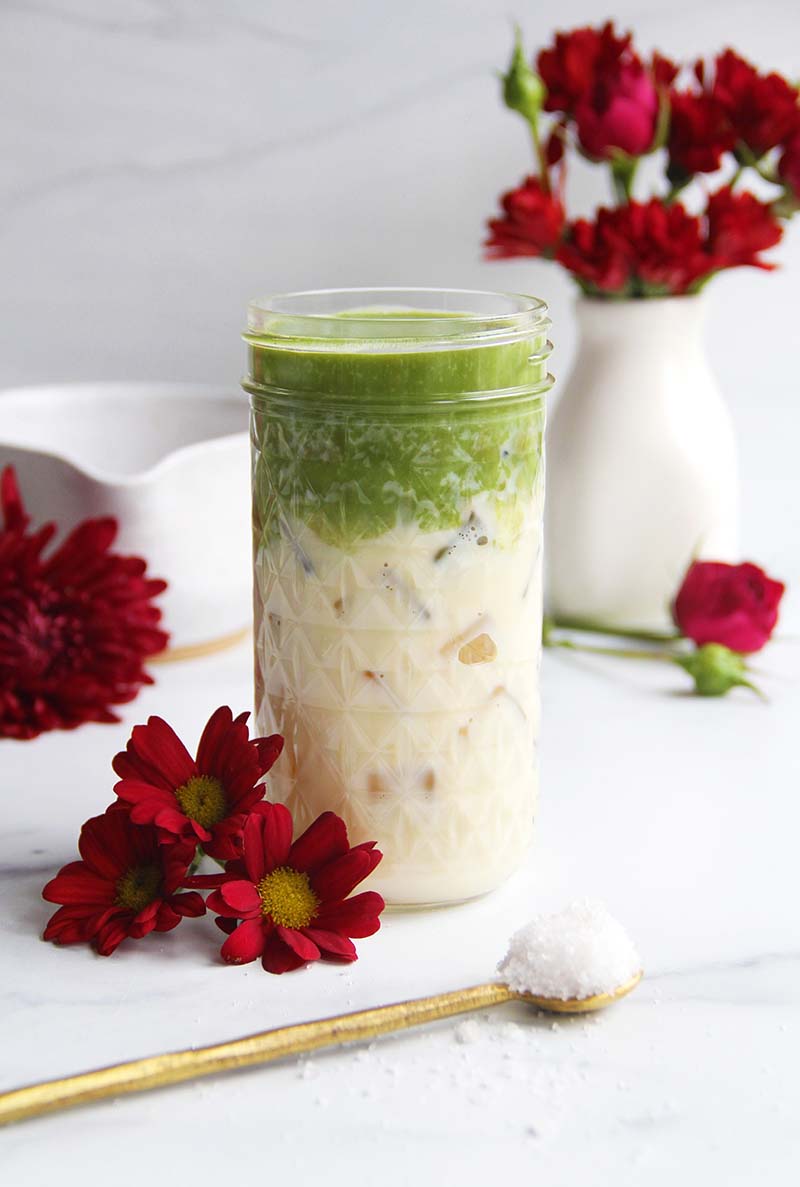 Homemade iced salted maple matcha latte in a glass jar, photographed in a bright white kitchen with red flowers and a matcha bowl.