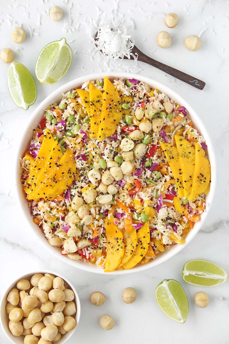 Overhead view of a tropical hearts of palm rice salad in a white serving dish on a marble countertop, garnished with macadamias and furikake, with lime wedges and extra macadamia nuts shown next to the salad.