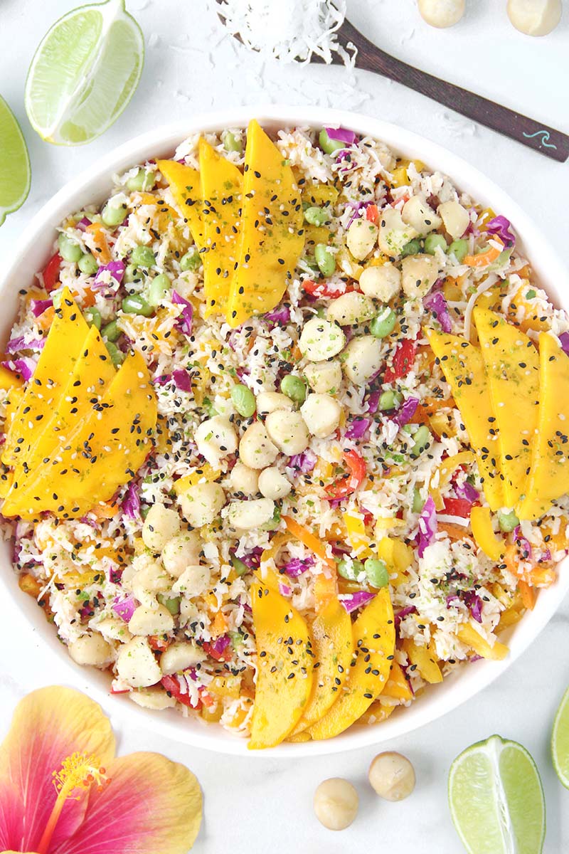 Tropical hearts of palm rice salad with colorful vegetables, mango, macadamias, and furikake, photographed overhead with lime wedges and hibiscus flowers on a marble surface.
