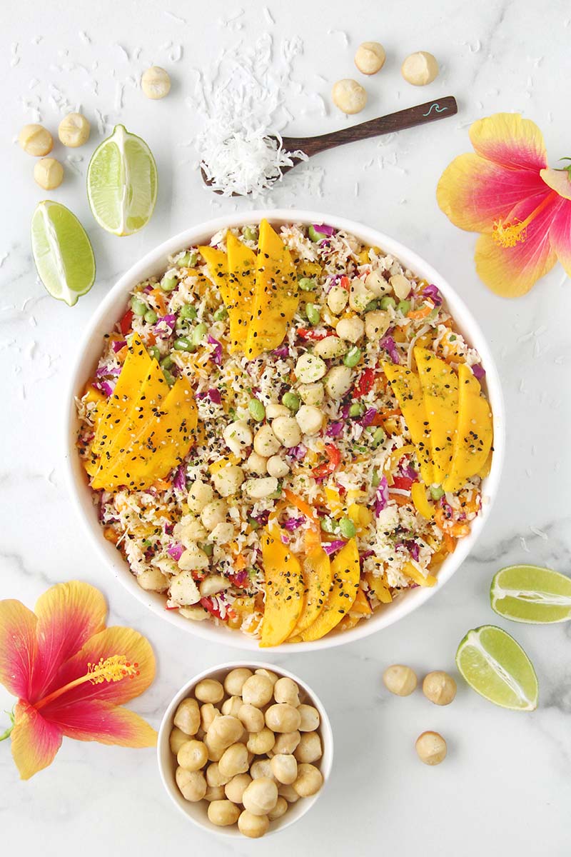 Overhead view of a tropical hearts of palm rice salad in a white bowl, garnished with macadamia nuts and furikake, shown with lime wedges, hibiscus flowers, and extra macadamias alongside.